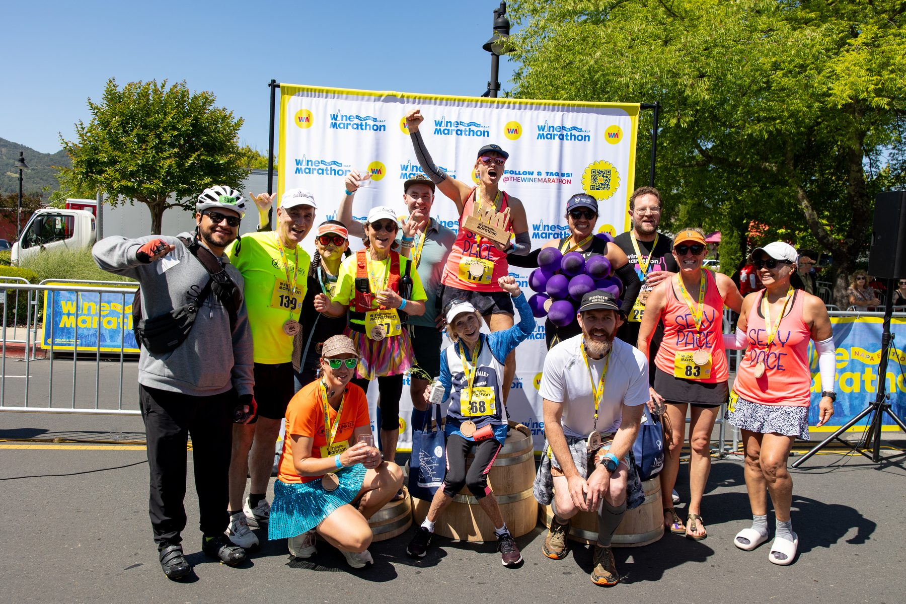 A large group of finishers posing with medals and wine glasses in front of the 2026 Wine Marathon backdrop in Napa Valley
